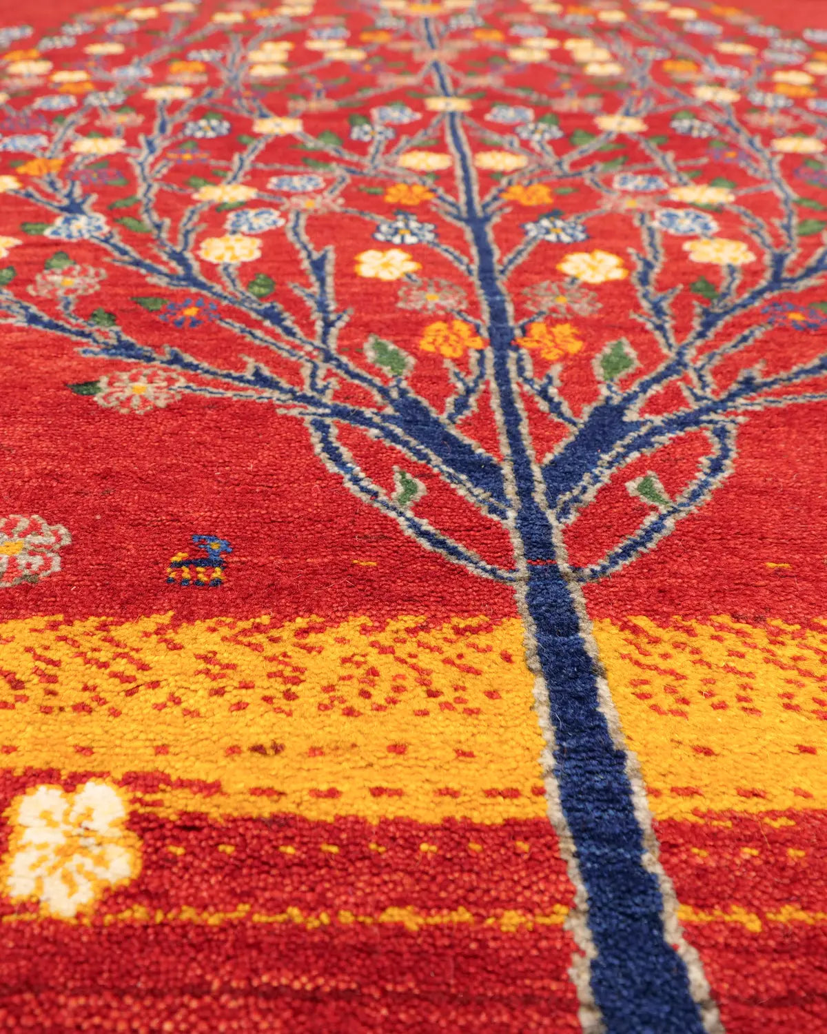 赤い背景に、青い幹と黄色や青の花が映える生命の樹デザインのギャッベ